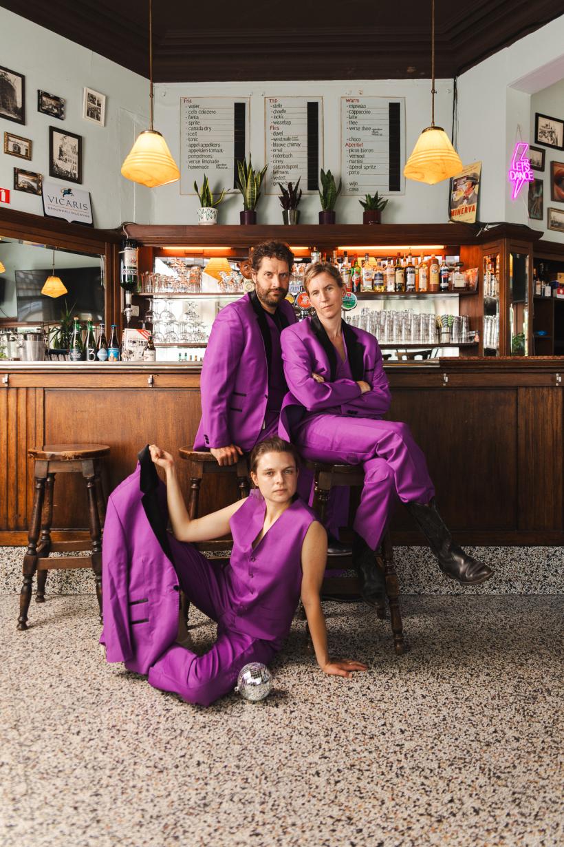 Drie personen (Suzanne Grotenhuis, Freek Vielen en Ellis Meeusen) in paarse pakken die aan een bar zitten.